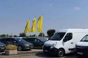 Auf dem Autohausgel&auml;nde in S&ouml;mmerda stehen mehrere Opel-Fahrzeuge, darunter ein wei&szlig;er Transporter und mehrere graue PKW. Im Hintergrund wehen drei gro&szlig;e gelbe Fahnen mit dem Opel-Logo. Der Himmel ist blau mit einigen leichten Wolken.