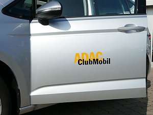 Nahaufnahme einer silbernen Autotür mit der Beschriftung ADAC ClubMobil in gelber und schwarzer Schrift. Im Bild ist der linke Außenspiegel, der Türgriff und ein Teil des Hinterrads zu sehen. Das Fahrzeug steht auf einem gepflasterten Platz.