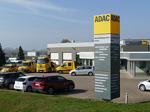 ADAC-Standort in Nordhausen; im Vordergrund eine große gelb-graue ADAC-Stele mit Beschriftung Mobilitätspartner Peter. Dahinter mehrere gelbe ADAC-Pannen- und Abschleppwagen und ein modernes, graues Gebäude mit Glasfassade und ADAC-Logo. Im Vordergrund parkende Autos, im Hintergrund Felder und Bäume bei klarem Himmel.