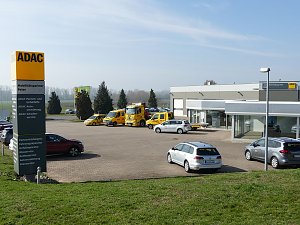 Alt-Text: Blick über das Gelände des ADAC Mobilitätspartners Peter in Nordhausen. Im Vordergrund ein großes gelbes ADAC-Schild mit Serviceleistungen, dahinter mehrere gelbe ADAC-Fahrzeuge und silberne Mietwagen, im Hintergrund das moderne Gebäudekomplex mit Glasfassaden und Werkstatt.