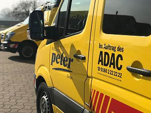 Gelber ADAC-Transporter vom Autohaus Peter, Nahaufnahme der Seitentür mit den Logos Autohaus Peter und Im Auftrag des ADAC sowie Telefonnummer. Im Hintergrund weitere gelbe ADAC-Fahrzeuge, gepflasterter Hof und Bäume.