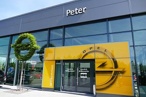 Eingang des Opel-Autohauses Peter in Nordhausen. Über der gläsernen Fassade steht der Schriftzug Peter. Vor dem Eingang befindet sich eine große gelbe Wand mit Opel-Logo und Herzlich Willkommen-Schriftzug. Links davon steht eine kunstvoll geschnittene grüne Hecke in Form des Opel-Logos. Der Eingangsbereich wirkt modern und einladend.