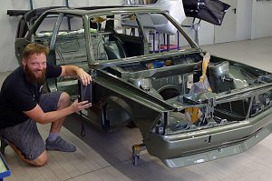 Tino Schwenzer kniet lächelnd neben einer teilweise montierten grünen Autokarosserie in einer Werkstatt. Er hält ein Messgerät an die Seitenwand, um den Farbton zu bestimmen. Im Hintergrund verschiedene Autoteile und Werkstattausrüstung.