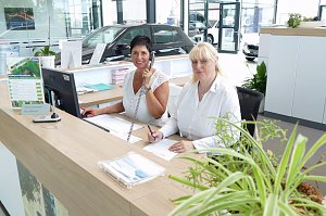 Zwei Serviceassistentinnen sitzen am Empfangstresen in einem modernen Autohaus. Eine Frau telefoniert und lächelt, die andere schreibt etwas auf. Beide tragen weiße Blusen. Auf dem Schreibtisch stehen ein Monitor, Unterlagen sowie Pflanzen. Im Hintergrund sind Autos und der helle Ausstellungsraum zu sehen.