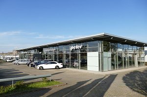 Autohaus Peter mit moderner Glasfassade und der Aufschrift �Gebrauchtwagen�. Vor dem Geb&auml;ude sind mehrere Mercedes-Fahrzeuge in verschiedenen Farben geparkt, darunter ein wei&szlig;er Wagen im Vordergrund. Weitl&auml;ufiger Parkplatz, blauer Himmel ohne Wolken.