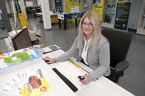 Junge Frau mit langen, blonden Haaren und Brille sitzt am Empfangsschalter des Autohauses. Sie trägt einen grauen Cardigan über einer weißen Bluse und lächelt leicht in die Kamera. Auf dem Schreibtisch liegen Prospekte, ein Monitor steht vor ihr. Im Hintergrund sind Verkaufs- und Beratungsbereiche des Opel-Autohauses sichtbar.