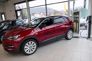 Roter Opel Grandland X Hybrid im Innenraum des Opel-Autohauses, während er an einer Ladestation angeschlossen ist. Das Fahrzeug steht vor großen Glasfenstern, durch die man Häuser und Hügel im Hintergrund sieht. Auf der Fahrerseite ist die Aufschrift Grandland X Hybrid sichtbar. Daneben weitere Fahrzeuge und ein hell gefliester Boden.