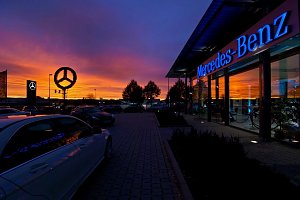 Mercedes-Benz Autohaus bei Sonnenuntergang. Der Himmel leuchtet in kräftigen Orange- und Lilatönen. Auf dem Parkplatz stehen mehrere Autos, der beleuchtete Mercedes-Stern und der Schriftzug �Mercedes-Benz� an der Glasfassade reflektieren das Licht.