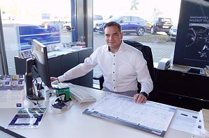 Autohaus-Mitarbeiter in weißem Hemd sitzt an einem Schreibtisch vor einem Computer, lächelt in die Kamera, Verkaufsunterlagen und Broschüren auf dem Tisch, Autos sind draußen durch das Fenster sichtbar.