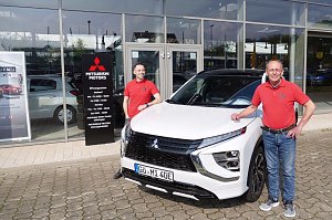 Zwei Mitarbeiter in roten Mitsubishi-Poloshirts stehen vor einem weißen Mitsubishi Eclipse Cross Plug-in Hybrid, vor dem Eingang des Autohauses Peter in Göttingen.