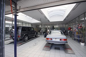 Helle Mercedes-Benz Werkstatt mit zwei Fahrzeugen auf Hebebühnen: links ein schwarzer Geländewagen (G-Klasse), rechts ein silberner Oldtimer. Im Hintergrund Glasbüros und Regale mit Autopflegeprodukten.