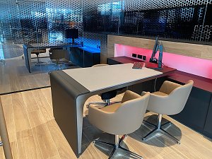 Modern eingerichtetes Verkäuferbüro im Autohaus mit hellem Holzboden, stilvollem Tisch, bequemen Stühlen und Computerarbeitsplatz. Im Hintergrund Glaswand mit Punktmuster.