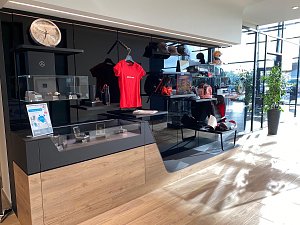 Mercedes-Benz Collection: Verkaufsbereich im Autohaus mit Verkaufsregalen, auf denen Mercedes-Fanartikel wie Shirts, Uhren und Mützen ausgestellt sind. Viel Tageslicht durch große Fenster.