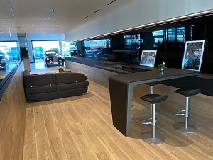 Moderner Kundenwartebereich im Autohaus mit Holzfußboden, dunkelbraunem Sofa, Hochtisch mit Barhockern, historischen Fotos und Blick auf ein klassisches Auto im Hintergrund.
