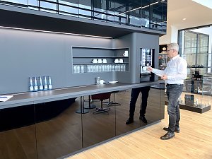 Mann im weißen Hemd zapft Kaffee am modernen Kaffeeautomaten in einer stilvollen Autohaus-Lounge mit Gläsern, Tassen und Wasserflaschen.