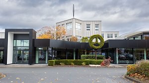 Frontansicht des modernen Mercedes-Autohauses in Nordhausen mit großen Glasflächen, schwarzer Fassade und einem Mercedes-Benz Logo als Formschnitt-Hecke, herbstliche Bäume im Hintergrund, bewölkter Himmel.