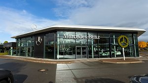 Modernes Mercedes-Benz Autohaus mit großer Glasfassade, Mercedes-Stern-Logo auf dem Dach und bepflanztem Mercedes-Symbol, blauer Himmel mit Wolken.