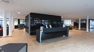 Moderner Empfangsbereich eines Mercedes-Benz Autohauses mit heller Holzoptik, schwarzem Empfangstresen, Glastrennwänden und Mercedes-Logo an der Wand.