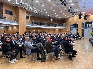 Weitwinkelaufnahme des Saals mit vielen Gästen, die aufmerksam nach vorn zur Bühne schauen.