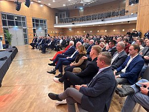 Publikum im Saal hört aufmerksam einer Rednerin auf der Bühne zu. Viele Männer im Anzug, einzelne Frauen im Publikum.