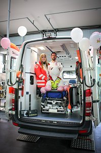 Ein geöffneter Rettungswagen steht im Autohaus. Zwei Mitarbeitende des Deutschen Roten Kreuzes stehen darin, vor ihnen sitzt ein großes Stoff-Einhorn auf einer Krankentrage. Rosa Luftballons schmücken den Wagen.