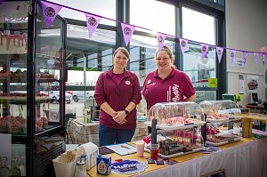 Zwei Frauen stehen hinter einem Kuchen- und Tortenstand im Autohaus. In einer Glasvitrine und auf Tischen sind Cupcakes und Torten ausgestellt. Rosa Flamingos dienen als Dekoration. Ein Banner mit dem Logo Happy Cakes hängt über dem Stand.