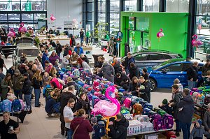 Großer Ausstellungsraum im �koda Autohaus, gefüllt mit vielen Menschen. In der Mitte sind lange Tische voller bunter Schulranzen. Mehrere �koda-Fahrzeuge stehen zwischen den Besuchern. Rosa Flamingo-Dekorationen hängen im Raum.