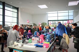 Viele Menschen, darunter Kinder mit Eltern, stehen an Tischen voller bunter Schulranzen im Autohaus. Rosa Flamingo-Dekorationen hängen von der Decke. Im Hintergrund sind Autos und große Fensterfronten sichtbar.
