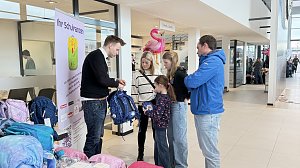 Ein Verkäufer zeigt einer Familie mit zwei Kindern einen blauen Schulranzen. Neben ihnen stehen weitere Ranzen. Im Hintergrund sind Flamingos und Schilder im Autohaus zu sehen.