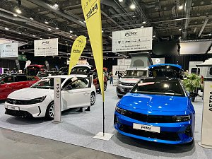 Vorne ein weißer Opel Corsa und daneben ein blauer Opel Astra, beide mit geöffneten Türen und Kofferräumen auf der Messe präsentiert.