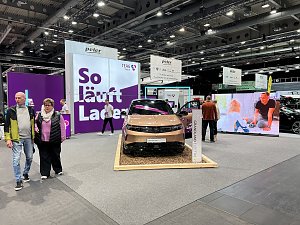 Ein goldfarbener Opel Grandland Electric auf einem Podest mit Holzunterlage, dahinter große lilafarbene Messewand mit Schriftzug So läuft Laden.