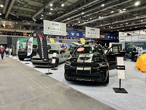 Ein schwarzer Dodge mit gelben Rallyestreifen vorne und ein grauer Dodge Challenger stehen nebeneinander. Daneben schwarze RAM-Fahrzeuge mit großen Bannern.