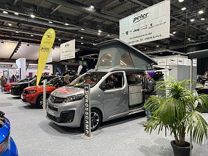 Grauer Opel Crosscamp mit geöffnetem Hochdach und Schiebetür, präsentiert auf der Messe. Daneben Besucher und weitere Opel-Fahrzeuge.