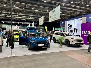 Ein blauer Peugeot und ein weißer Peugeot SUV nebeneinander auf der Ausstellungsfläche, im Hintergrund große Messebanner und lilafarbene Wand So läuft Laden.