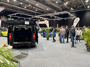 Schwarzer Transporter mit geöffneter Heckklappe, ausgestellt auf der Messe. Besucher laufen durch die Halle, viele Fahrzeuge im Hintergrund.
