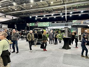 Besucher gehen zwischen verschiedenen Ständen hindurch. Sicht auf SUVs und Camper, mit schwarzen und grünen Messebannern im Hintergrund.