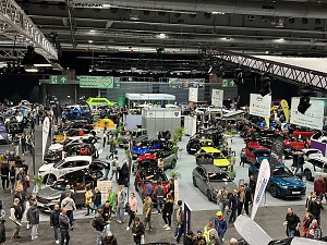 Panoramaaufnahme der Messehalle mit zahlreichen Autos in verschiedenen Farben, vielen Besuchern und großen Markenbannern.