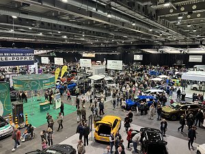 Breiter Überblick über die Automobilmesse mit dichten Besuchergruppen, Fahrzeugen aller Marken und farbigen Ausstellungsflächen.