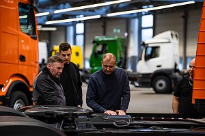 Mehrere M&auml;nner inspizieren technische Details am Fahrgestell eines orangefarbenen Mercedes-Benz eActros in einer Produktionshalle.
