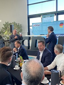 Helmut Peter begrüßt mit Mikrofon die Gäste in der Autowelt Peter. Mehrere Personen sitzen an runden Tischen mit Getränken und Frühstücksgeschirr. Im Hintergrund große CDU-Buchstaben und eine Bühne mit Team Merz 2025-Plakat.