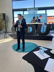 David Gregosz im Anzug spricht vor Publikum in einer Autohaus-Halle. Hinter ihm Bühne mit CDU-Logo und Podiumsteilnehmern, vorne auf dem Tisch Broschüren und eine Tasse.