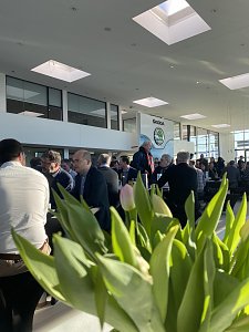 Innenaufnahme des Autohauses mit vielen Gästen, die an schwarzen Tischen sitzen. Im Vordergrund unscharfe Tulpen, im Hintergrund große Fenster und das Skoda-Logo.