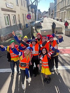 Gruppe von als Narren verkleideten Menschen in orangefarbenen Warnwesten steht auf einer Straße und winkt fröhlich in die Kamera.