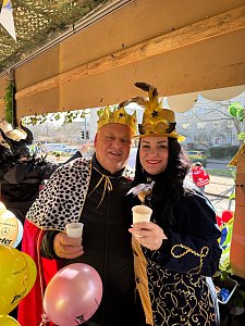 Helmut Peter und Christina Dost, verkleidet mit goldenen Kronen und prächtigen Umhängen, halten jeweils einen weißen Becher und lächeln in die Kamera. Bunte Luftballons im Vordergrund.