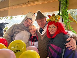 "Vier Personen in farbenfrohen Kostümen � darunter ein Bärenkostüm, ein Dirndl und ein Fantasiekostüm mit roter Perücke � stehen beieinander und lächeln in die Kamera, umgeben von bunten Luftballons.