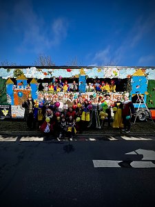 Große Gruppe verkleideter Menschen posiert vor und auf einem bunt gestalteten Karnevalswagen mit Märchenmotiven, viele halten bunte Luftballons.
