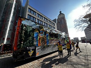 Seitliche Ansicht eines bunt dekorierten Karnevalswagens mit Märchenmotiven, gezogen von einem orangefarbenen LKW, im Hintergrund ein historisches Gebäude mit Uhrturm, mehrere als Narren verkleidete Personen laufen daneben.