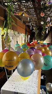 "Reihe bunter Luftballons mit der Aufschrift �Helau, Alaaf & Ahoi!� und dem Autohaus-Peter-Logo, befestigt an weißen Stäben, unter einer mit Pflanzen und Sternen dekorierten Decke.