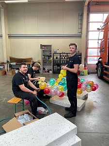 Drei Männer in schwarzer Arbeitskleidung sitzen und stehen in einer Werkstatt, binden bunte Autohaus-Peter-Luftballons zusammen, im Hintergrund ein orangefarbener LKW.