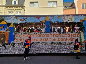 Seitenansicht des bunt gestalteten Karnevalswagens mit dem Schriftzug �In jedem Märchen nah und fern fährt er stolz den Silberstern�, mehrere verkleidete Menschen stehen darauf, zwei Personen in Narrenkostümen davor.
